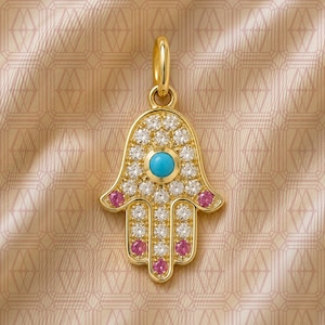 May include: A gold Hamsa pendant featuring a central round turquoise stone, surrounded by clear pavé-set stones. The hand-shaped charm includes four small pink stones at the fingertips and base, finished with a gold bail at the top for hanging.