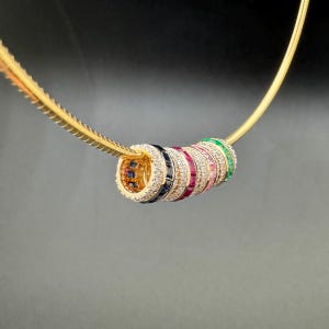 May include: A gold chain necklace with a pendant made of stacked rings. Each ring is set with a row of small gemstones in a different color, including green, blue, pink, and red.