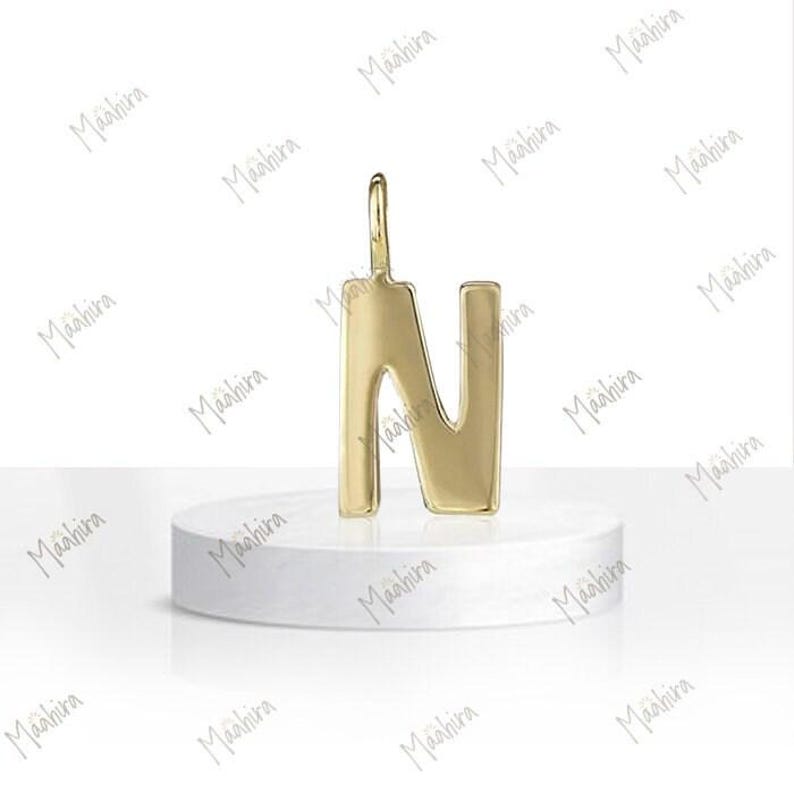 May include: A polished gold-tone metal pendant shaped like the capital letter N. The letter features a smooth, reflective surface and a small vertical loop at the top for attachment. The design is simple, clean, and uniform in its metallic finish.