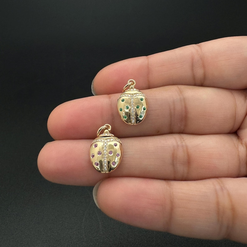 May include: Two gold ladybug charms with clear and colored gemstones. One charm has green gemstones and the other has pink gemstones.