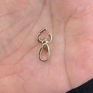 May include: A small gold-toned metal lobster claw clasp featuring a swivel mechanism at the top and a spring-loaded gate on the lower loop. The hardware has a polished, reflective finish and is designed for attaching charms or jewelry components.