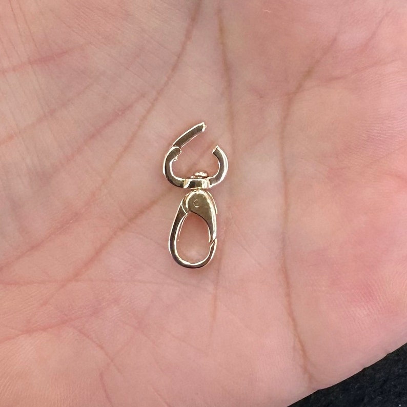 May include: A small gold-toned metal lobster claw clasp featuring a swivel mechanism at the top and a spring-loaded gate on the lower loop. The hardware has a polished, reflective finish and is designed for attaching charms or jewelry components.