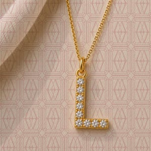 May include: A gold-toned necklace featuring a pendant shaped like the capital letter L. The pendant is encrusted with a series of small, clear, round-cut stones along its entire surface, suspended from a delicate matching gold-toned chain.