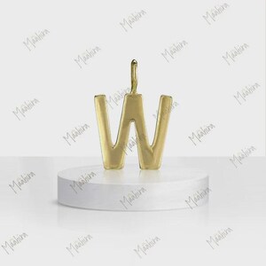 May include: A gold-toned metal pendant in the shape of the capital letter W. The pendant features a smooth, polished surface and a small vertical loop at the top center for attachment. It is displayed standing upright on a white circular base.