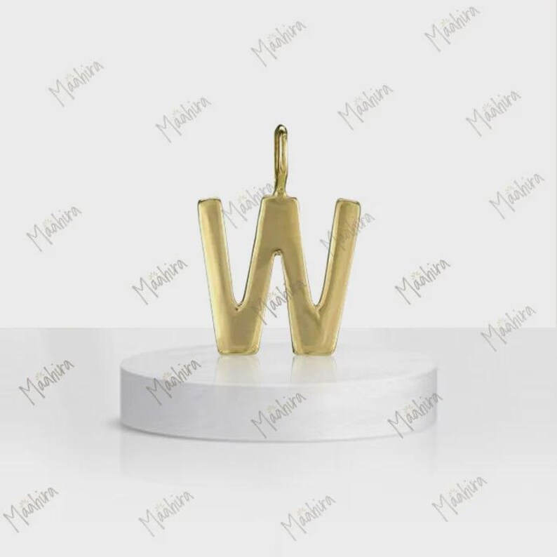 May include: A gold-toned metal pendant in the shape of the capital letter W. The pendant features a smooth, polished surface and a small vertical loop at the top center for attachment. It is displayed standing upright on a white circular base.