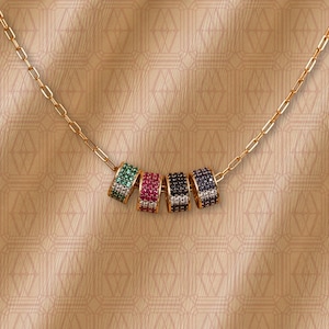 May include: A gold-toned chain necklace featuring four cylindrical beads, each encrusted with small pavé stones in green, red, black, and dark blue. The beads are arranged horizontally along the center of the delicate gold-colored metal chain.