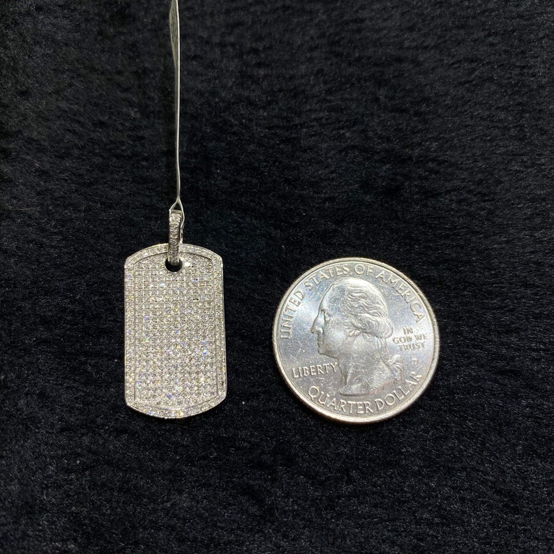 May include: A silver dog tag pendant with a pave setting of small diamonds. The pendant is shown next to a US quarter for size comparison.