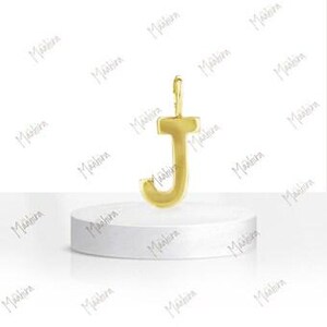 May include: A gold-colored metal pendant in the shape of the capital letter J. The letter features a smooth, polished surface with a small vertical loop at the top for attachment. The pendant is displayed standing upright on a white circular base.