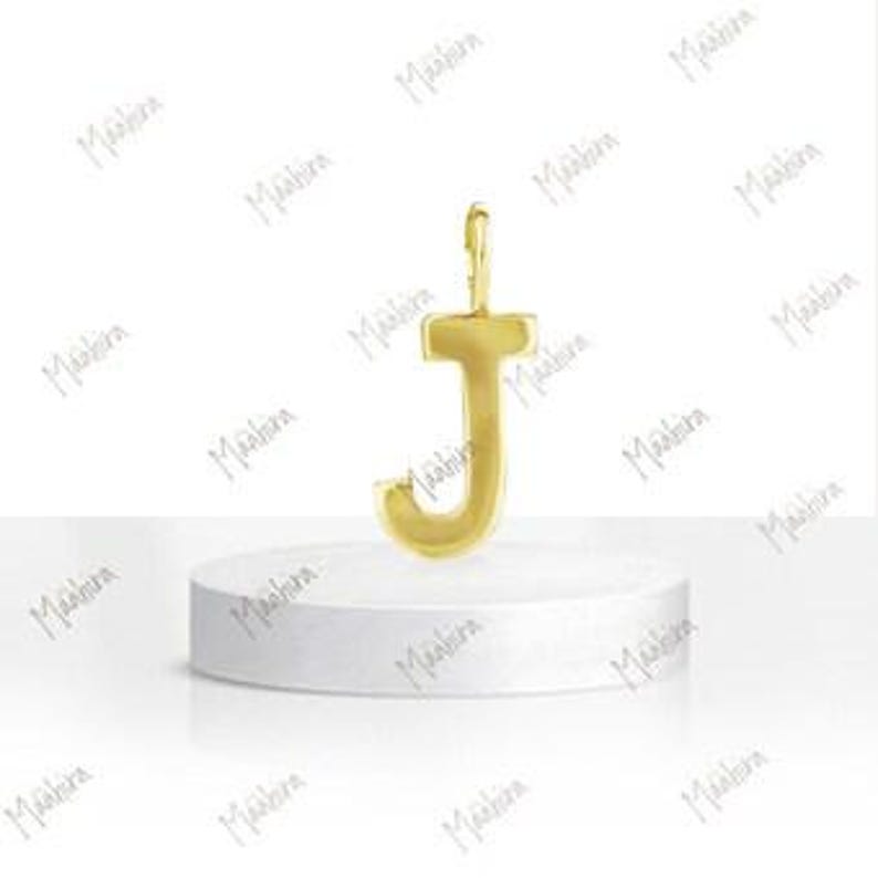 May include: A gold-colored metal pendant in the shape of the capital letter J. The letter features a smooth, polished surface with a small vertical loop at the top for attachment. The pendant is displayed standing upright on a white circular base.