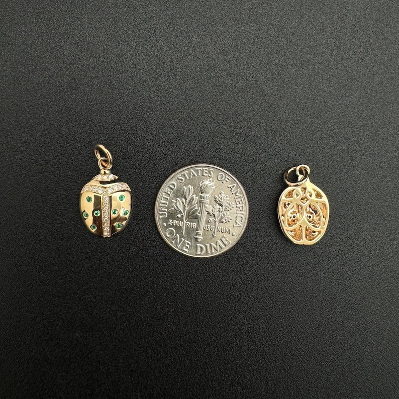May include: Two gold charms, one shaped like a ladybug with green and white stones, the other a more abstract design, shown next to a US dime for scale.