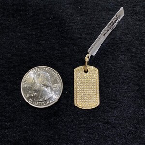 May include: A gold dog tag pendant with a pave setting of small clear stones. The pendant is next to a US quarter for size comparison.