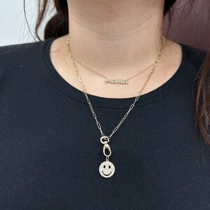 May include: A layered gold-tone necklace set featuring two distinct chains. The shorter chain displays a script-style pendant reading mama, while the longer paperclip-style chain features a circular smiley face charm with clear stone accents.