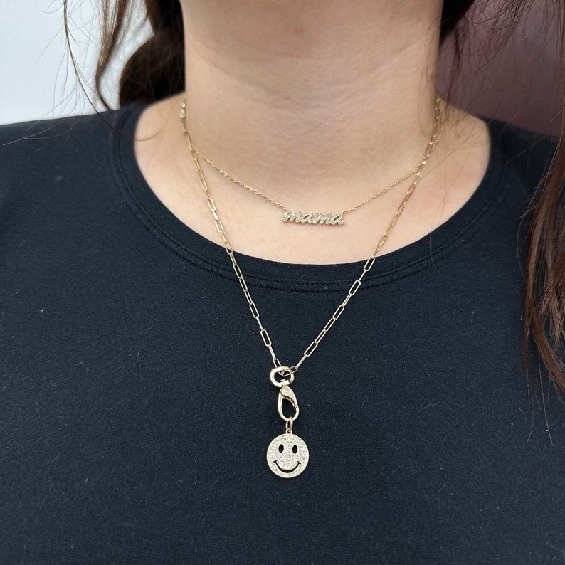 May include: A layered gold-tone necklace set featuring two distinct chains. The shorter chain displays a script-style pendant reading mama, while the longer paperclip-style chain features a circular smiley face charm with clear stone accents.