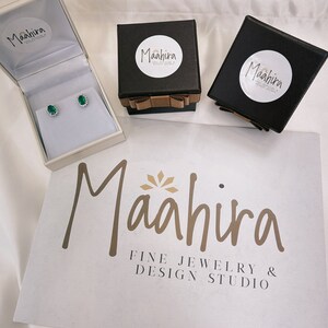 May include: A pair of oval-cut green gemstone stud earrings with a halo of small clear stones, presented in an open cream-colored box. Two black square gift boxes with brown ribbon bows and Maahira branding sit nearby on a white surface.