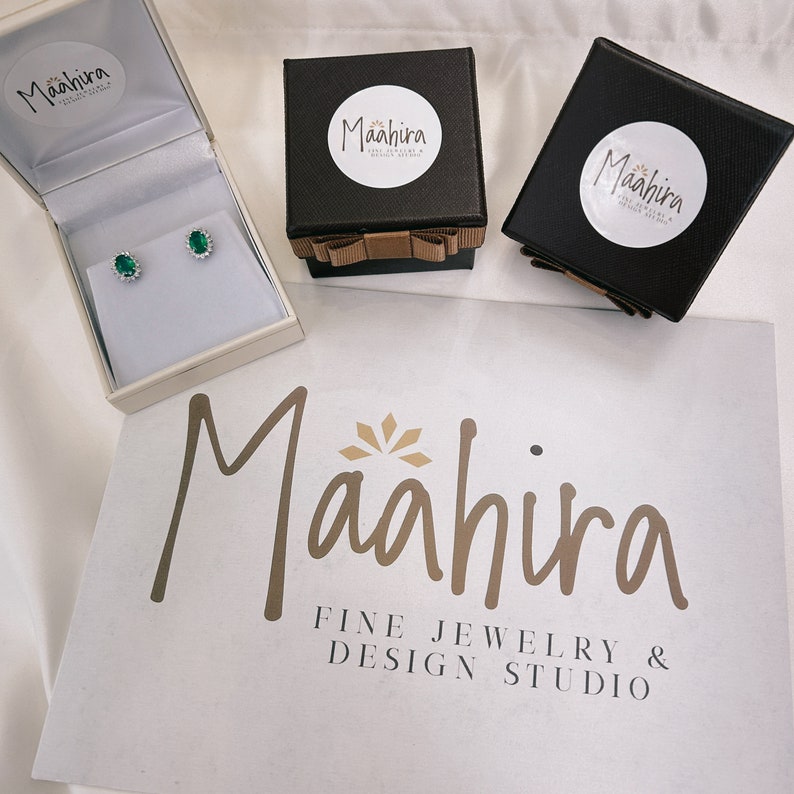 May include: A pair of oval-cut green gemstone stud earrings with a halo of small clear stones, presented in an open cream-colored box. Two black square gift boxes with brown ribbon bows and Maahira branding sit nearby on a white surface.
