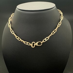 May include: A gold-toned chain necklace featuring elongated rectangular links with a central horizontal bar in each link. The necklace is secured with a lobster claw clasp and a small oval jump ring, creating a uniform and repeating geometric pattern.