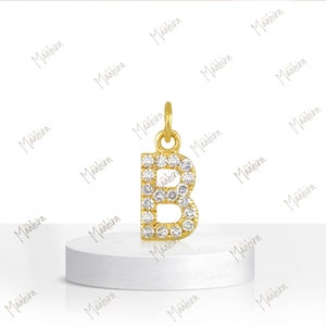 May include: A gold-toned letter B pendant featuring a series of small, clear, round-cut stones embedded along the surface of the letter. The pendant includes a small circular jump ring at the top for attachment to a chain or cord.
