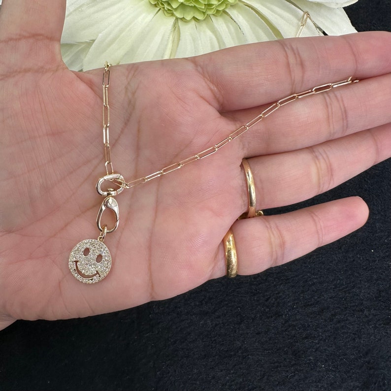 May include: A gold-toned chain necklace featuring a paperclip link design and a circular pendant shaped like a smiley face. The smiley face pendant is covered in small, clear, sparkling stones and is attached to the chain with a lobster clasp.