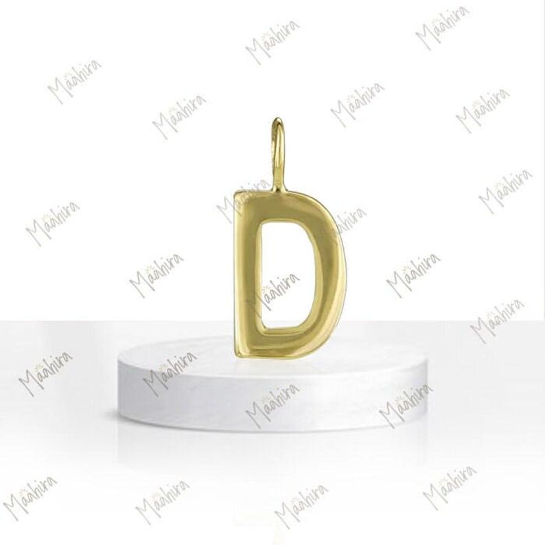 May include: A polished gold-tone pendant in the shape of the capital letter D. The pendant features a smooth, rounded surface and includes a small vertical loop at the top for attachment to a chain or cord, presented as a single jewelry piece.