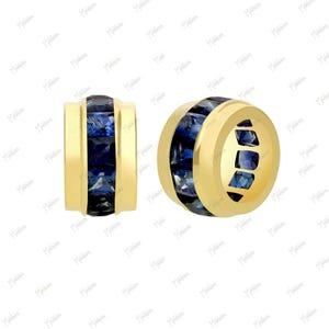 May include: Two cylindrical gold beads featuring a central channel set with square-cut deep blue sapphires. The gold bands frame the stones on both sides, creating a smooth, polished finish on the exterior of these jewelry components.