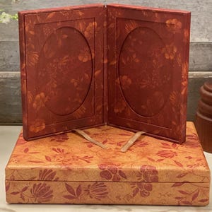 Puede incluir: Un marco de fotos rojo con estampado floral y dos aberturas ovaladas, apoyado sobre una caja a juego. El marco y la caja tienen una estética vintage. Un barril de mantequilla de madera y un jarrón con flores están en el fondo.