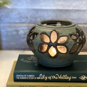May include: A teal ceramic candle holder with flower cutouts, illuminated by a warm glow. It sits atop two vintage books, "Lily of the Valley" and "The Fir Tree." A vase of flowers is in the background.
