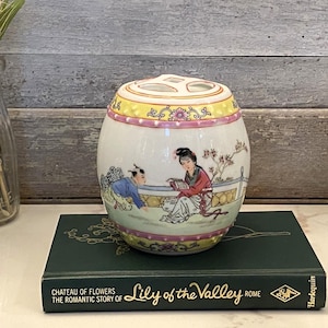 Puede incluir: Un portacepillos de cerámica ovalado con tapa. El portacepillos está decorado con una escena de figuras con vestimenta tradicional, con detalles en amarillo, rosa y verde. Se encuentra sobre un libro verde titulado "Lily of the Valley".