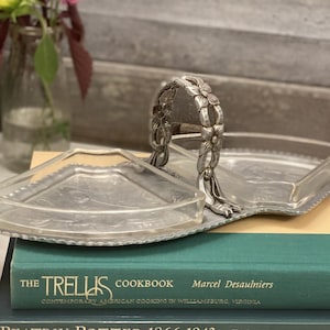 Op de afbeelding: Een zilverkleurige serveerschaal met een handvat versierd met bloemmotieven. De schaal heeft twee compartimenten en een heldere glazen inzet. Het staat op een stapel boeken, waaronder "The Trellis Cookbook".