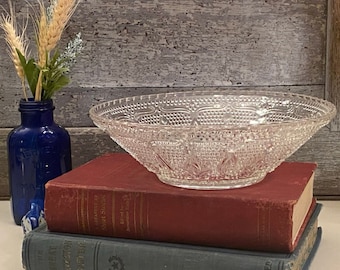 May include: A clear glass bowl with a decorative pattern sits on top of two vintage books. The books are red and blue. A small blue glass bottle with dried wheat stalks is in the background.