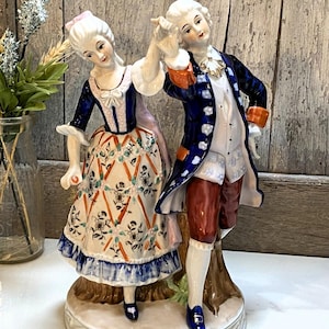 May include: A porcelain figurine depicting a couple in 18th-century attire dancing. The figure on the left wears a floral patterned skirt with a blue bodice, while the figure on the right wears a blue coat, white vest, and reddish-brown breeches.