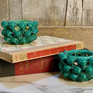 May include: Two teal ceramic candle holders with a textured, overlapping petal design. One holder sits atop a stack of two hardcover books, while the second holder rests on the surface next to the books. The books have red and cream spines.