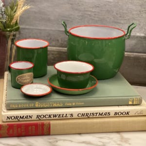 May include: A collection of green enamelware with red trim, including a pot with handles, cups, and a small dish. The items are arranged on a stack of vintage books, creating a nostalgic, rustic aesthetic.