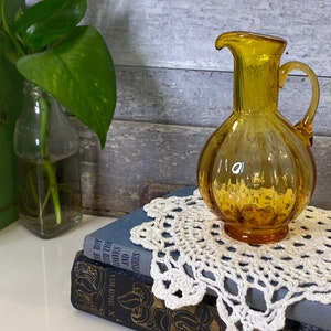 May include: A small amber glass pitcher with a rounded body and a handle. The pitcher is sitting on a white crocheted doily on top of a stack of books.