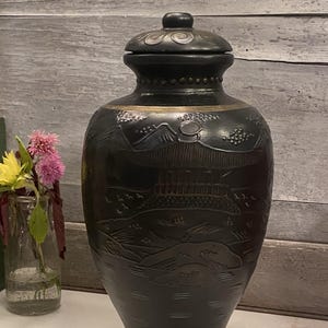 Op de afbeelding: Een donkere, decoratieve vaas met een deksel, met een reliëf landschapsontwerp. De vaas is zwart met gouden accenten en staat op een wit oppervlak. Naast de vaas staat een kleine glazen vaas met roze en gele bloemen.