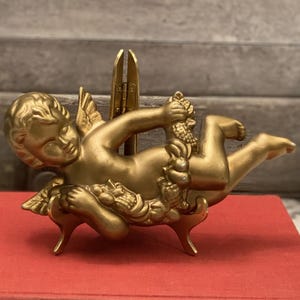 Puede incluir: Una figura de querubín dorada con los brazos y las alas extendidas descansa sobre un libro rojo. El querubín sostiene una guirnalda de uvas. El libro tiene letras doradas y está junto a un jarrón con flores.