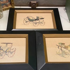 May include: Three framed prints depicting antique carriages. Each print has a beige background with a detailed illustration of a different carriage design. The frames are black with a gold inner border, creating a vintage aesthetic.