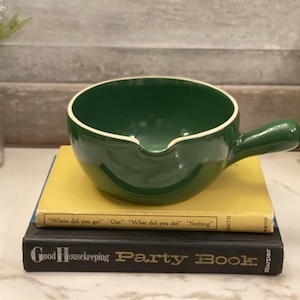 May include: A green ceramic soup bowl with a handle, resting on two books. The bowl has a white rim and a spout. The books are yellow and black, with text visible on the spines. A vase with flowers is in the background.