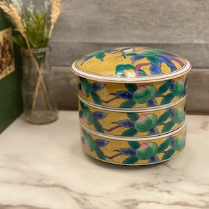 May include: A three-tiered, round ceramic container with a yellow background and a floral design. The container has a lid and is decorated with green leaves, pink fruit, and blue accents. The container is on a white surface.