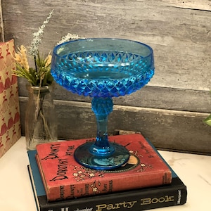 Puede incluir: Una vibrante copa de vidrio azul vintage con un patrón de diamantes en el cuenco y el tallo. La copa se encuentra sobre una pila de libros antiguos, incluido uno titulado "Dorothy Dainty at the Shore". El fondo presenta una pared de madera rústica y una pequeña planta.