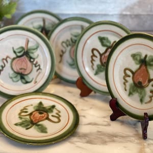 May include: A set of six ceramic plates with a hand-painted pear design. Each plate features a white center with a brown and green pear illustration, a green border, and a brown accent ring. The plates are displayed on a marble surface.