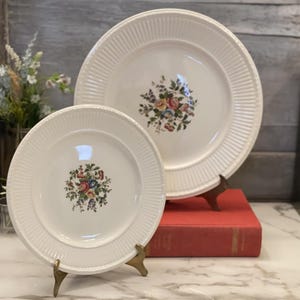 以下が含まれることがあります： 中央に花柄模様のある2つの白いセラミックプレート。プレートはリブ付きのエッジで、真鍮製のスタンドに展示されています。花柄のデザインには、ピンク、イエロー、ブルー、グリーンが含まれています。プレートは赤い本の上にあります。