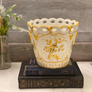 May include: A white ceramic planter with yellow floral designs and a scalloped rim. The planter sits atop two antique books with gold lettering. A small glass vase with greenery is in the background.