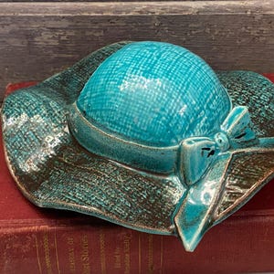 May include: A ceramic hat-shaped trinket dish in turquoise and brown. The hat features a textured crown and brim, with a bow detail. The dish is resting on a red book.