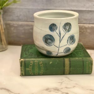 May include: A small, white ceramic pot with blue leaf designs sits atop a vintage green book. The pot has a rounded shape and a simple, elegant design. The book is titled "American Household Adviser."