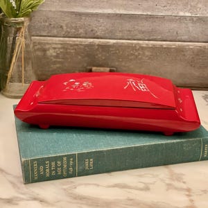 May include: A glossy red rectangular box with rounded edges, resting on a teal book. The box has a decorative design and text on the lid. The book is titled "Manners and Morals in the Age of Optimism".