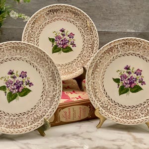 Puede incluir: Tres platos decorativos vintage con fondo crema y borde de filigrana dorada. Cada plato presenta un racimo de violetas moradas con hojas verdes. Los platos se exhiben sobre soportes dorados.