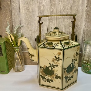 May include: A hexagonal ceramic teapot with a cream-colored base and dark green floral patterns. The teapot features a matching lid and a prominent brass-colored metal swing handle attached to the top. The design includes painted botanical motifs.