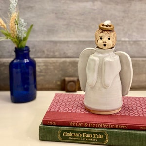 May include: A ceramic angel figurine with a white robe and wings, a tan face, and a gold halo. The angel is standing on a stack of books, with a blue glass bottle holding flowers in the background.