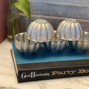 May include: Four silver metal vintage jello molds on a blue and black book with the text "Good Housekeeping Party Book".