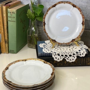 May include: Set of four white ceramic plates with a gold and brown floral pattern around the edge. The plates have a scalloped edge and are stacked on top of each other.